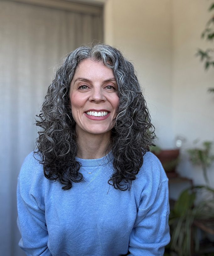 Smiling woman with curly gray hair in a blue sweater indoors.