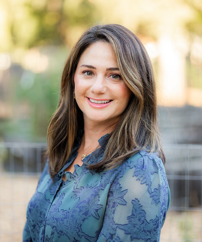 Smiling woman with shoulder-length hair outdoors in a blue patterned top.