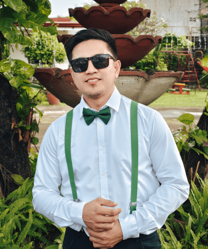 Man in white shirt with green suspenders and bow tie outdoors.