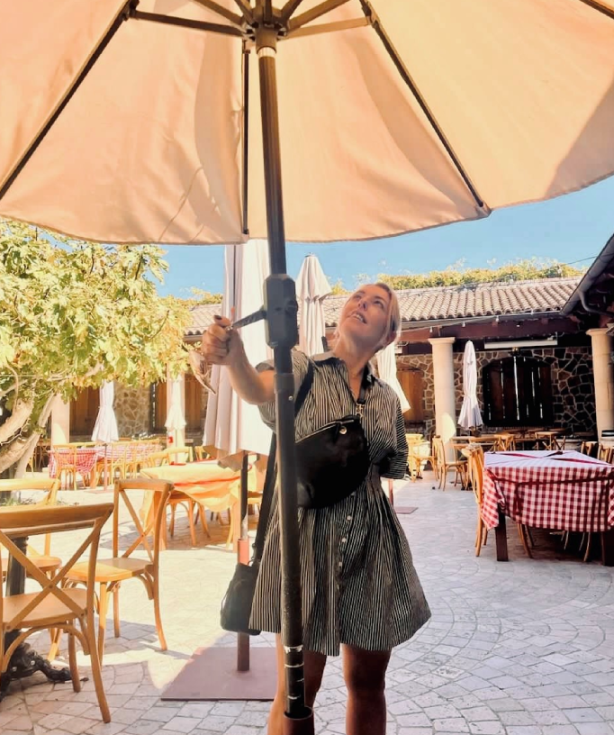 Person smiling while holding a large umbrella in an outdoor cafe.